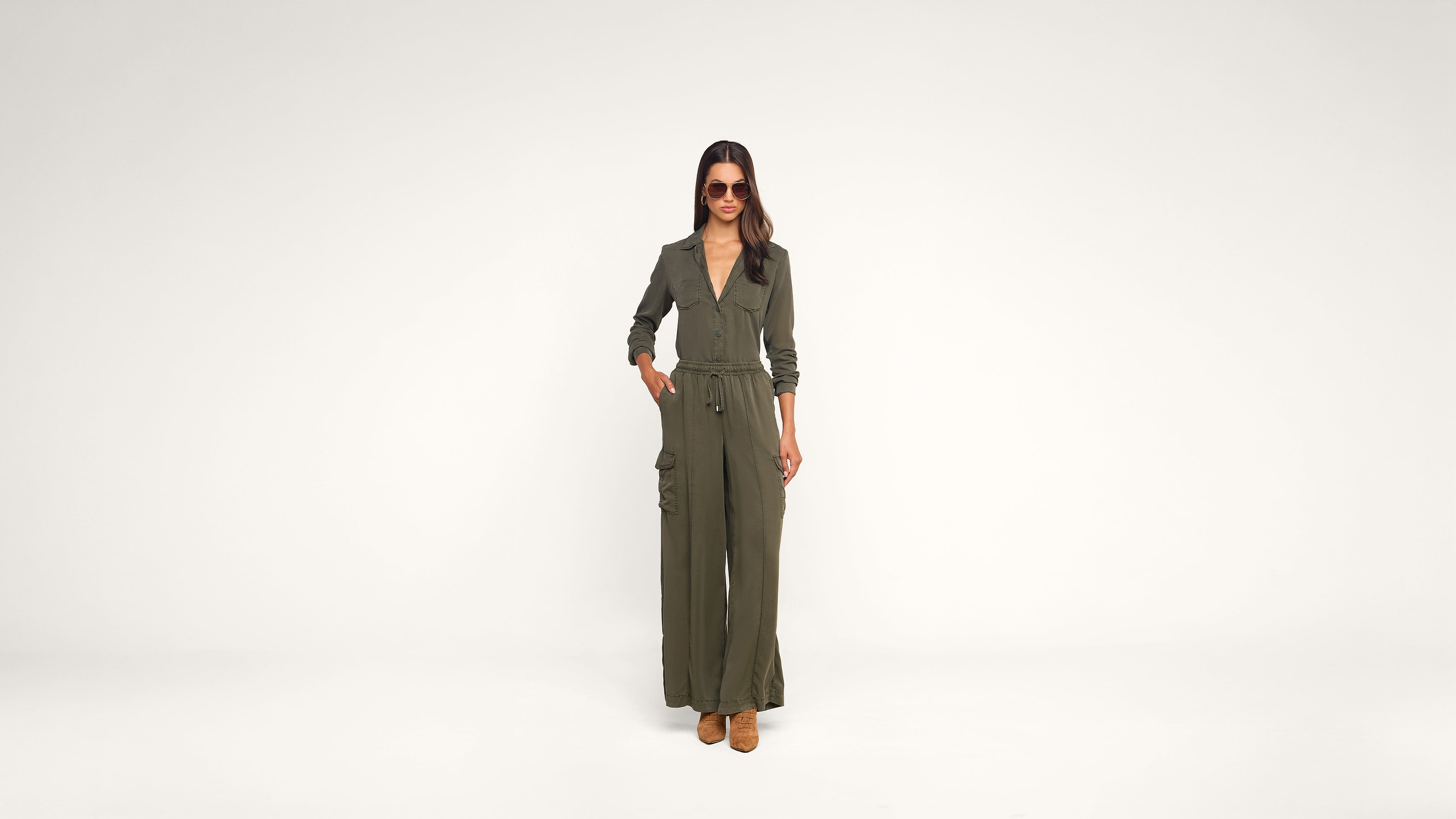 A woman wearing sunglasses and an olive green jumpsuit stands confidently with one hand in her pocket against a plain white background.