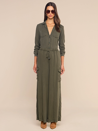 A woman stands facing forward, wearing sunglasses and the Isla Shirt—an olive green, long-sleeve maxi dress crafted from featherweight twill with a cinched waist and front pockets. She also wears brown shoes. The background is beige and plain.