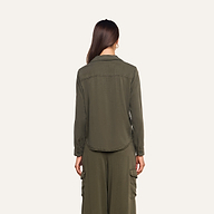 A woman stands facing away from the camera, wearing the olive green Isla Shirt paired with matching cargo pants featuring pockets. She has long brown hair and is set against a plain, light background.