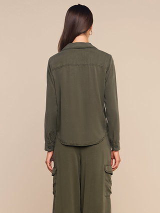 A woman stands with her back to the camera, wearing the olive green Isla Shirt in featherweight twill along with matching wide-leg pants featuring side pockets, set against a plain beige background.