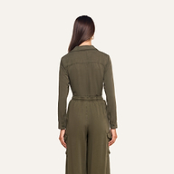 A woman with long brown hair stands facing away, wearing the Isla Shirt in olive green featherweight twill, featuring an elastic waistband and cargo pockets, against a plain light background.
