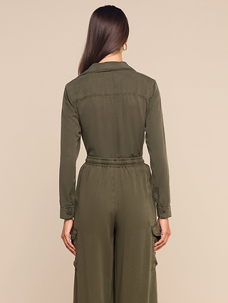 A woman with long brown hair, wearing the Isla Shirt in olive green featherweight twill, stands with her back to the camera against a plain beige background.