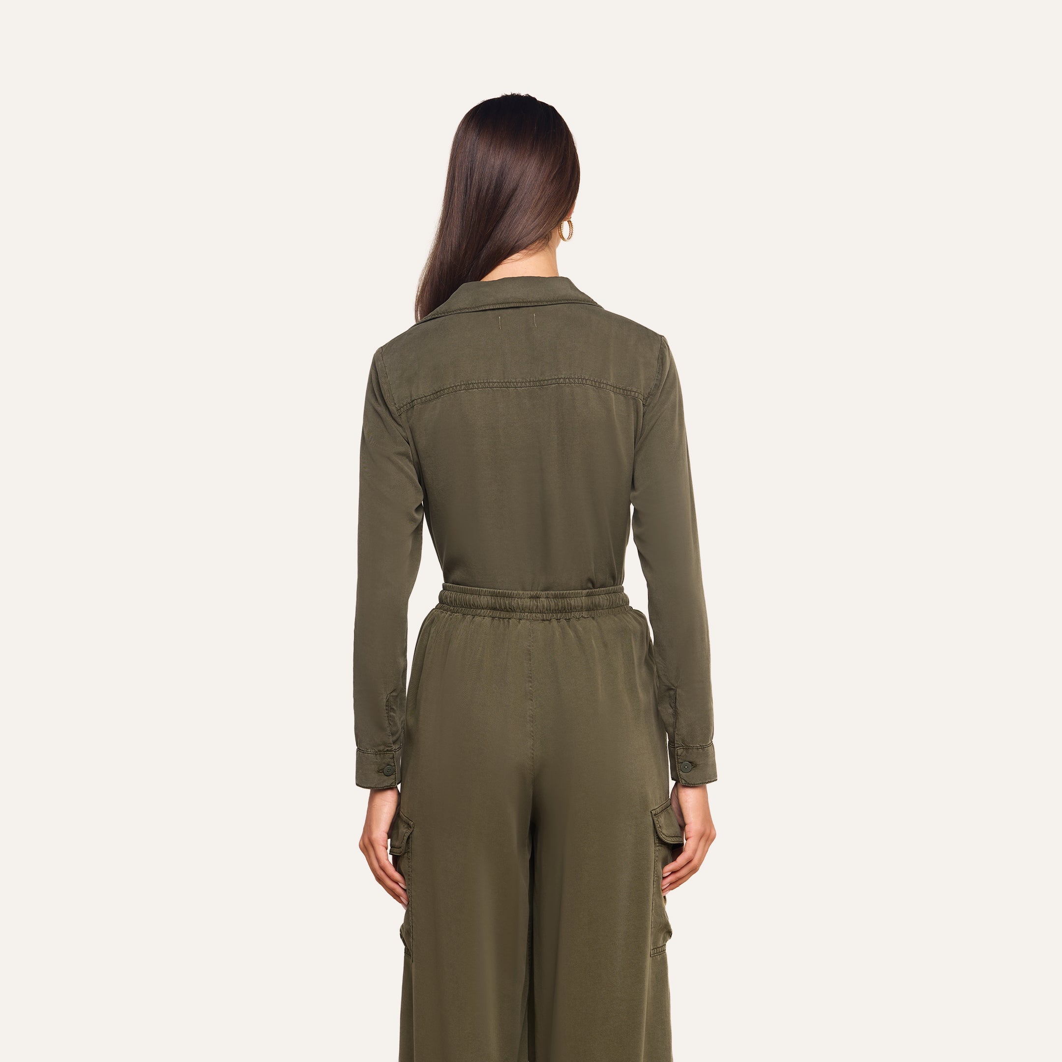 A woman with long brown hair stands facing away, wearing the Isla Shirt in olive green featherweight twill, featuring an elastic waistband and cargo pockets, against a plain light background.