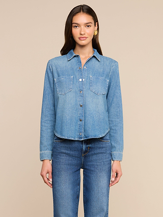 A woman stands against a beige background, wearing the Isla Shirt—a medium wash chambray top with two front pockets—paired with matching denim jeans. She has long brown hair and looks directly at the camera with a neutral expression.