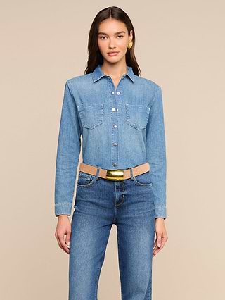 A person wearing the Isla Shirt—a long-sleeve medium wash chambray shirt—tucked into high-waisted blue jeans and a beige belt stands facing forward against a plain light background.