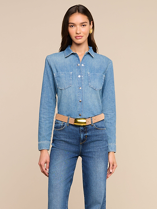 Woman with long brown hair wearing the Isla Shirt—a featherweight denim button-up—paired with medium wash blue jeans and a beige belt with a gold buckle, standing against a plain beige background and looking at the camera with a neutral expression.