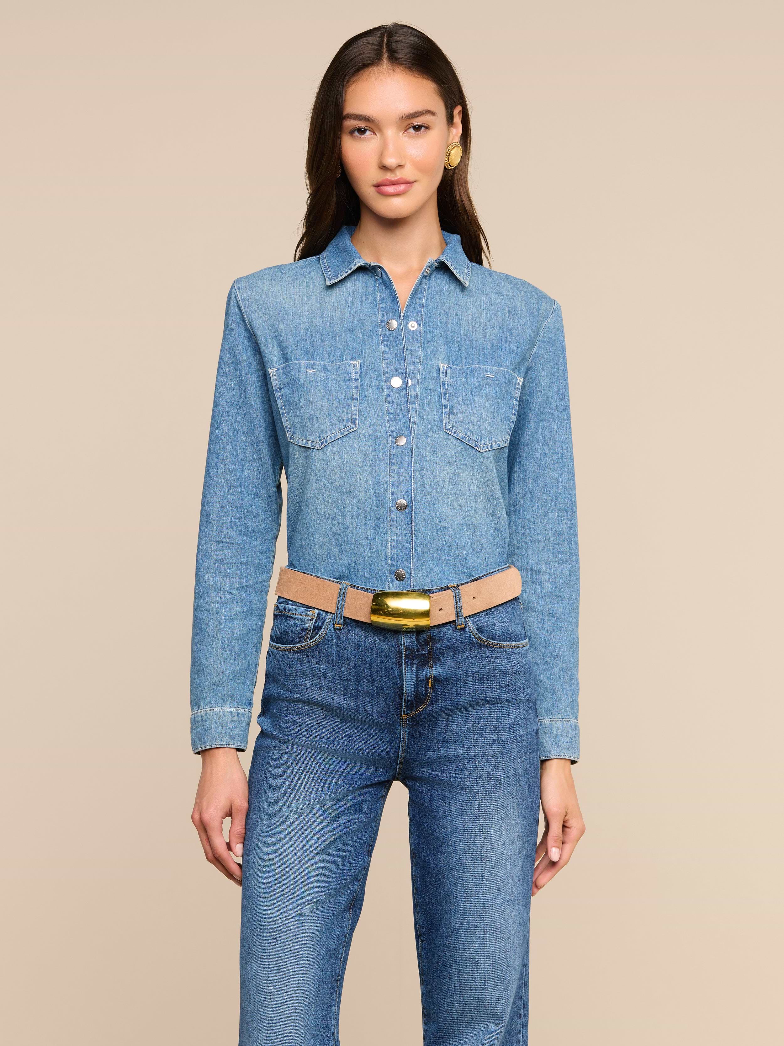 A person wearing the Isla Shirt—a long-sleeve medium wash chambray shirt—tucked into high-waisted blue jeans and a beige belt stands facing forward against a plain light background.