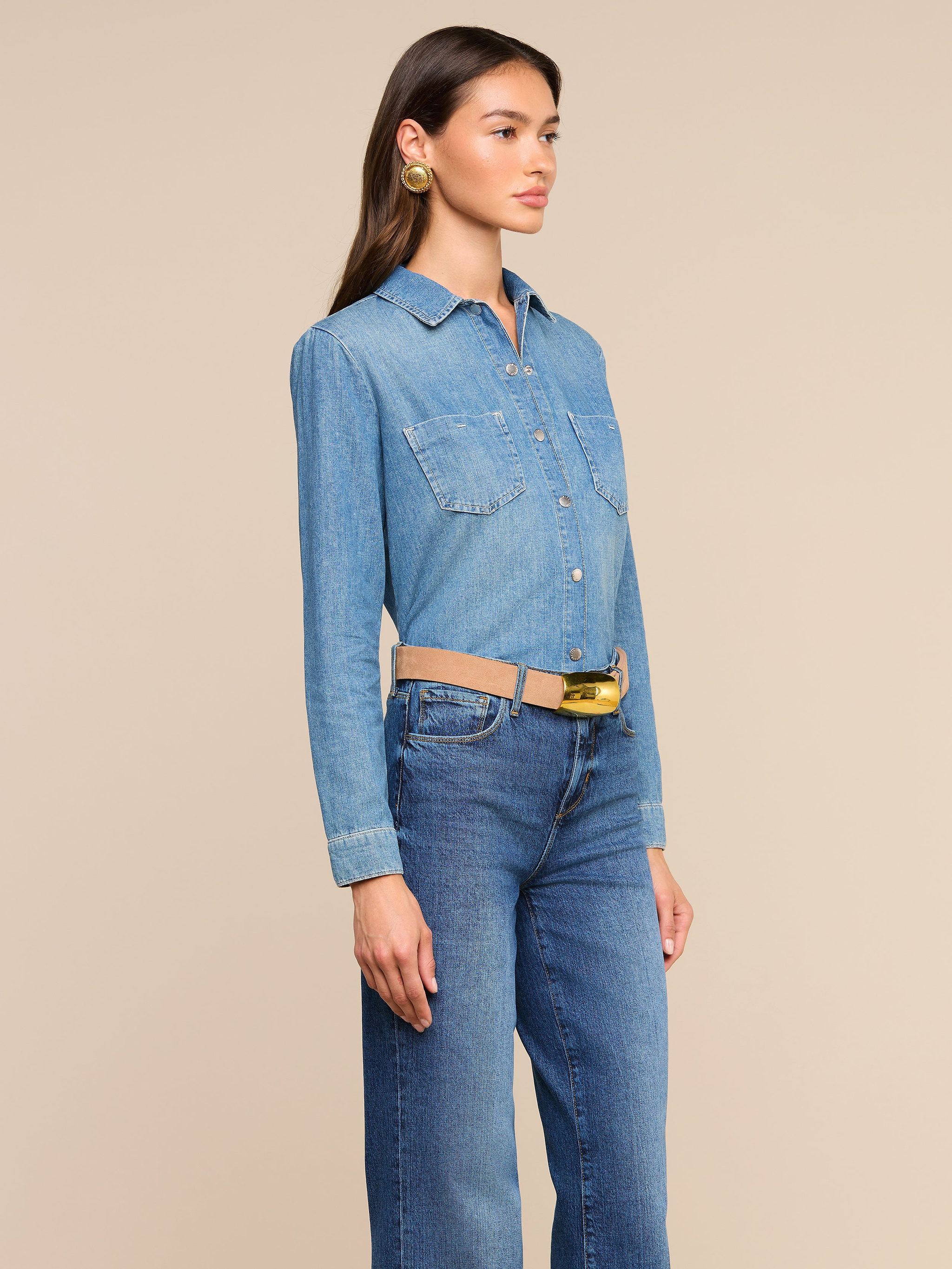 Woman standing and facing right, wearing the Isla Shirt in featherweight denim chambray paired with matching medium wash blue jeans, accessorized with a beige belt and large round gold earrings, against a plain off-white background.