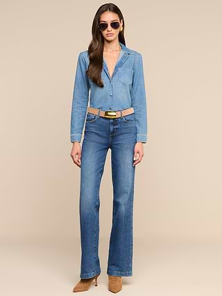 A woman wears the Isla Shirt, a medium wash chambray shirt, tucked into blue jeans with a tan belt and brown heeled shoes, accessorized with sunglasses, standing against a plain white background.