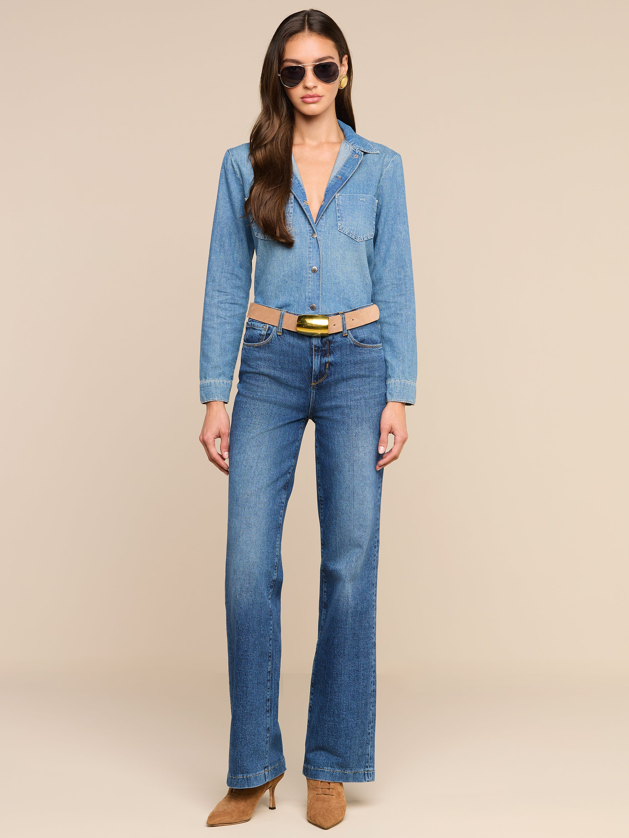 A woman wears the Isla Shirt, a medium wash chambray shirt, tucked into blue jeans with a tan belt and brown heeled shoes, accessorized with sunglasses, standing against a plain white background.