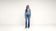 Woman standing in a minimal studio wearing a denim jacket and flared jeans, a brown belt, brown shoes, and sunglasses. She has long brown hair and a neutral expression.