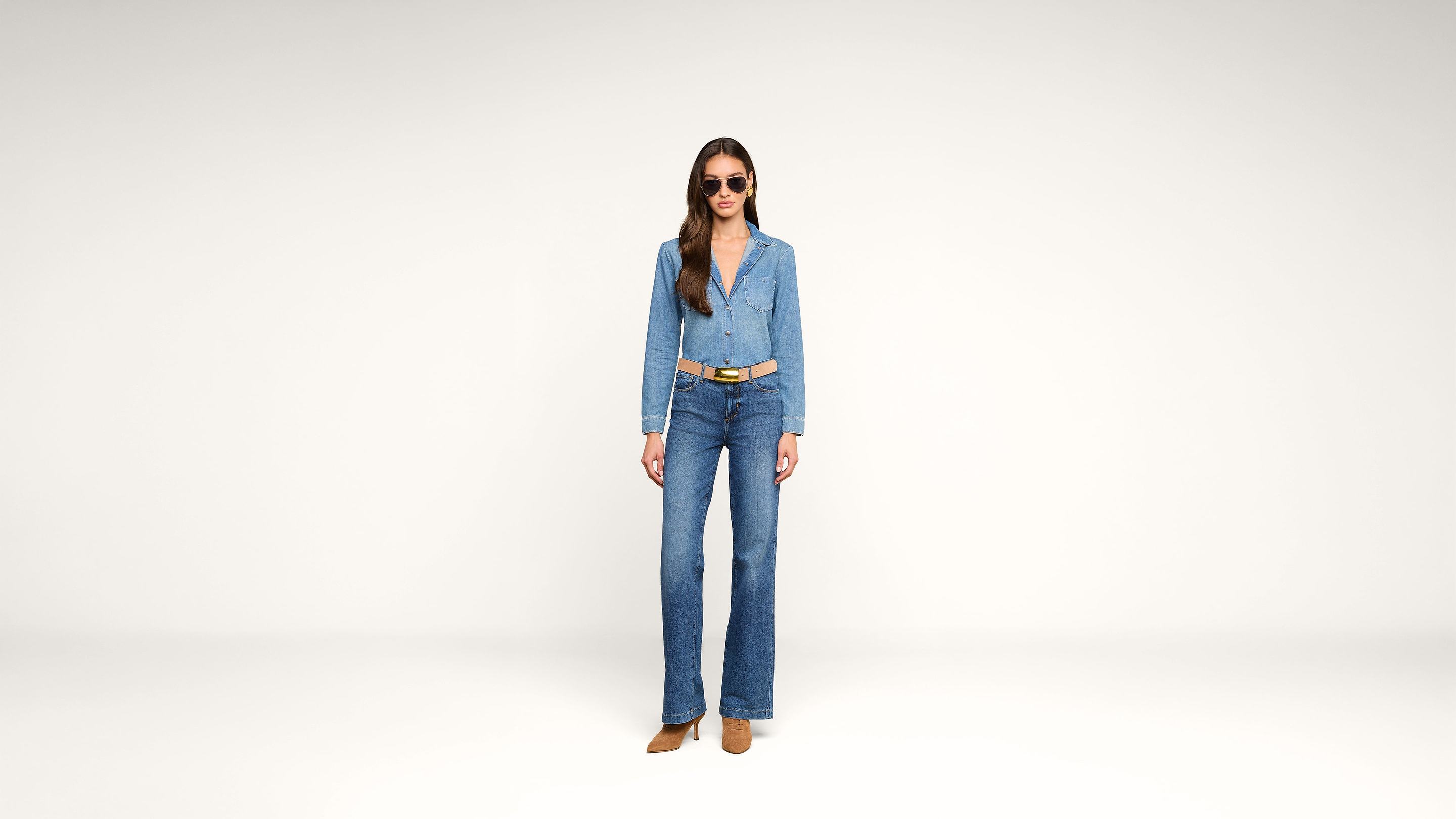 Woman standing in a minimal studio wearing a denim jacket and flared jeans, a brown belt, brown shoes, and sunglasses. She has long brown hair and a neutral expression.