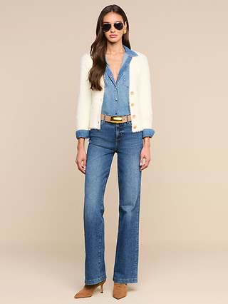 Woman wearing sunglasses, a white cardigan over the Isla Shirt tucked into blue high-waisted jeans, a black belt, and tan pointed-toe heels, standing against a plain white background.