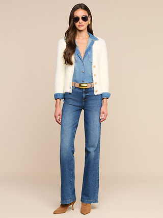 A woman wearing sunglasses, a white cardigan, the Isla Shirt, and medium-wash jeans with a brown belt and tan heeled boots stands against a beige background.