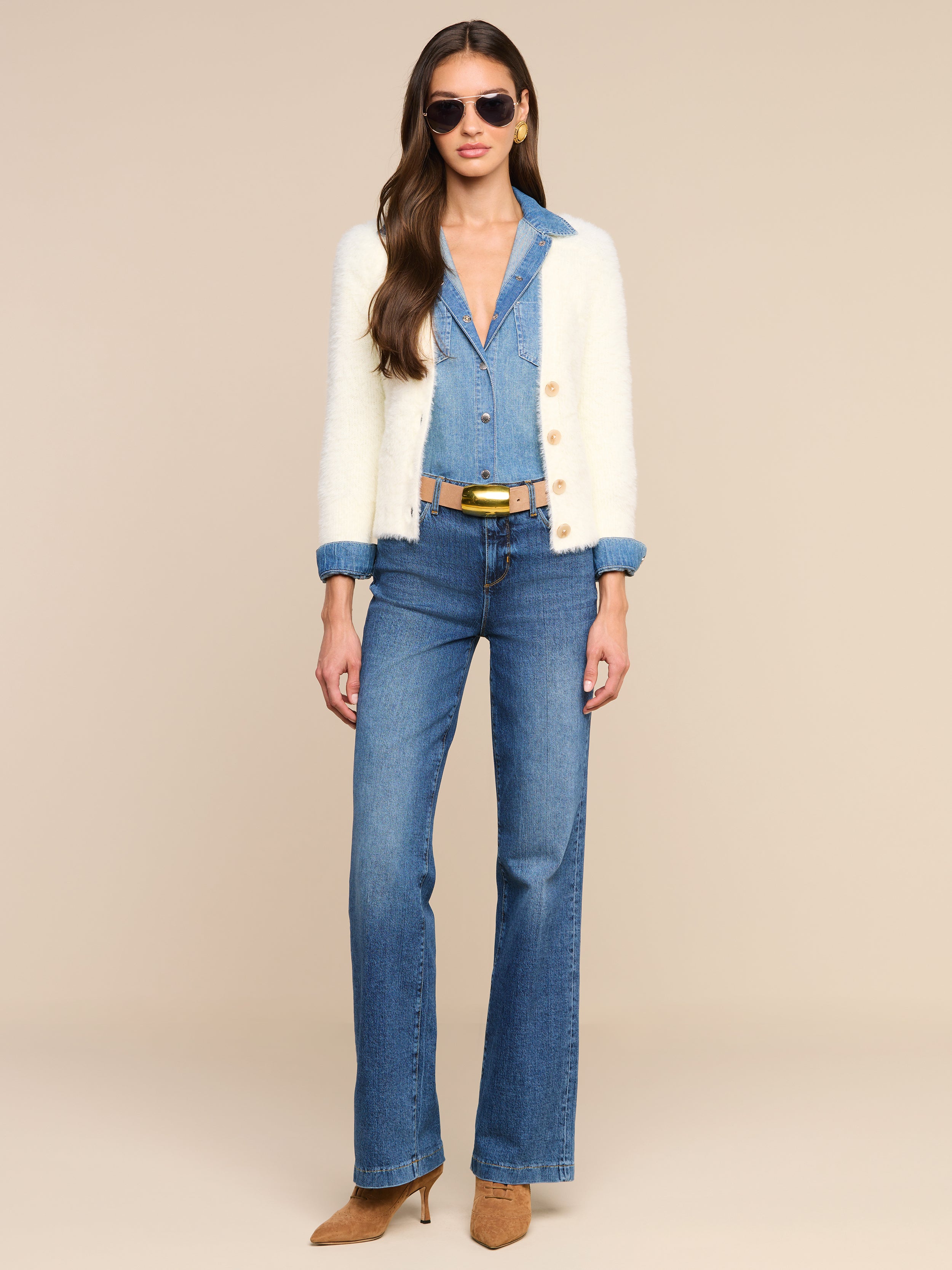 Woman wearing sunglasses, a white cardigan over the Isla Shirt tucked into blue high-waisted jeans, a black belt, and tan pointed-toe heels, standing against a plain white background.