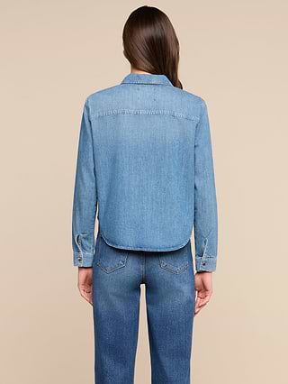 A person with long brown hair stands facing away, wearing the Isla Shirt and blue jeans against a plain light background.