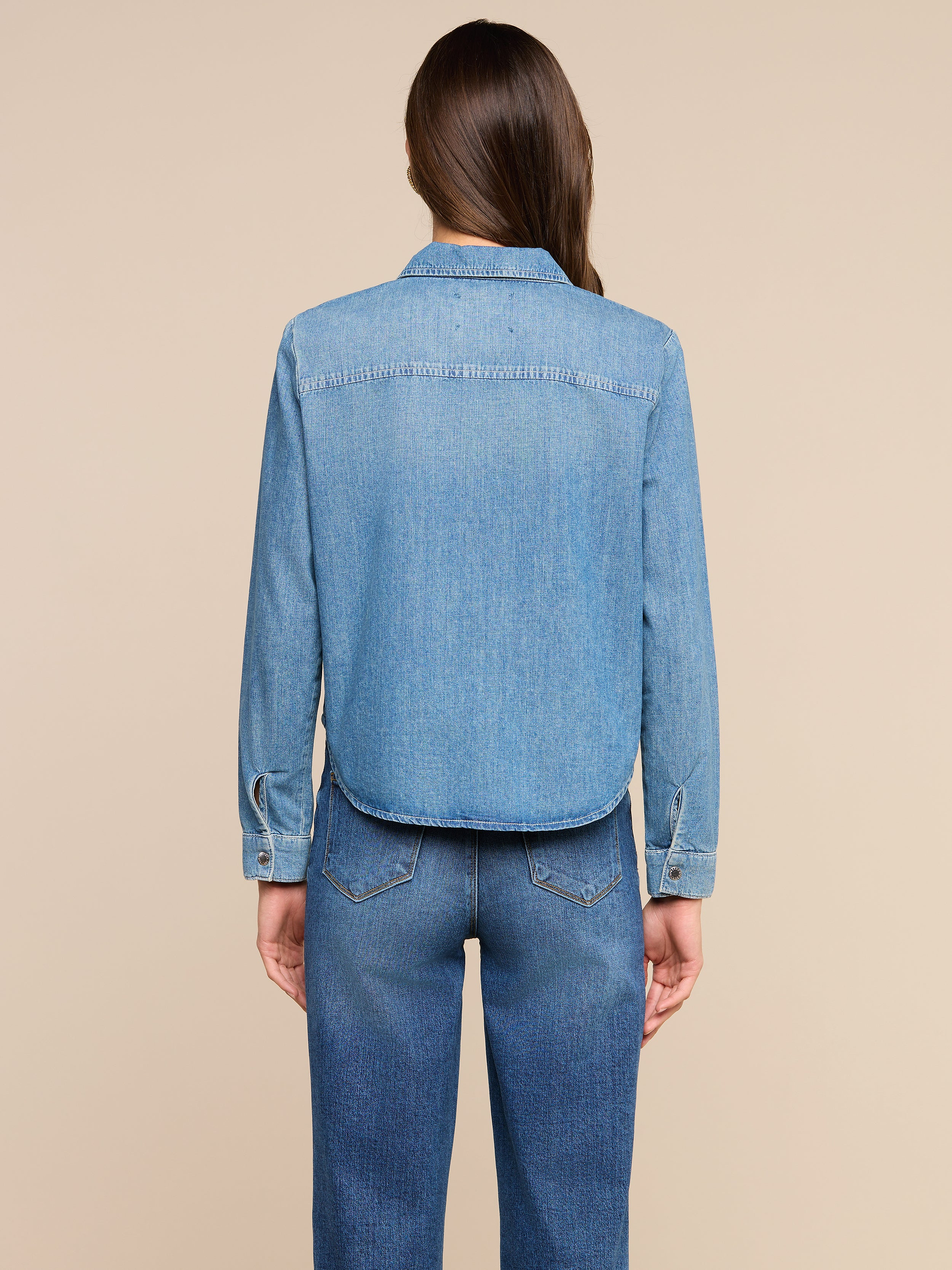 A person with long brown hair stands facing away, wearing the Isla Shirt and blue jeans against a plain light background.
