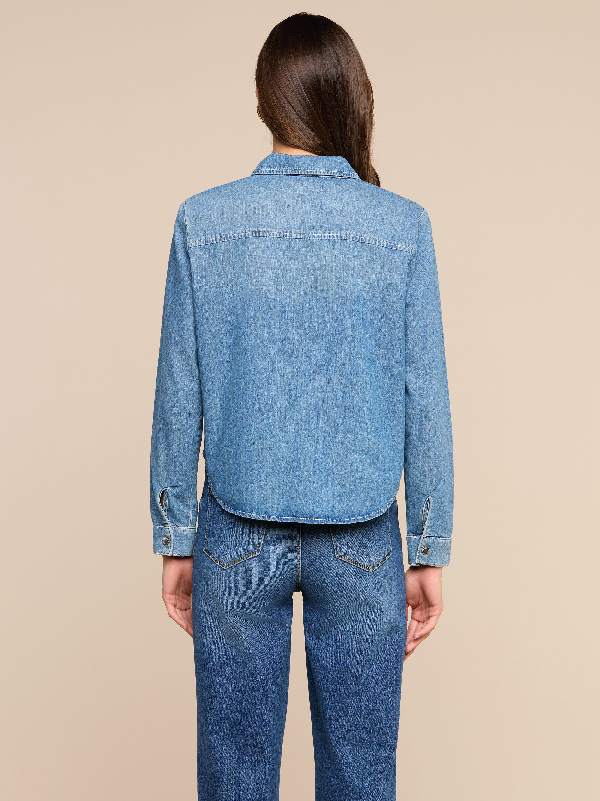 A person with long brown hair stands facing away, wearing the Isla Shirt and blue jeans against a plain light background.