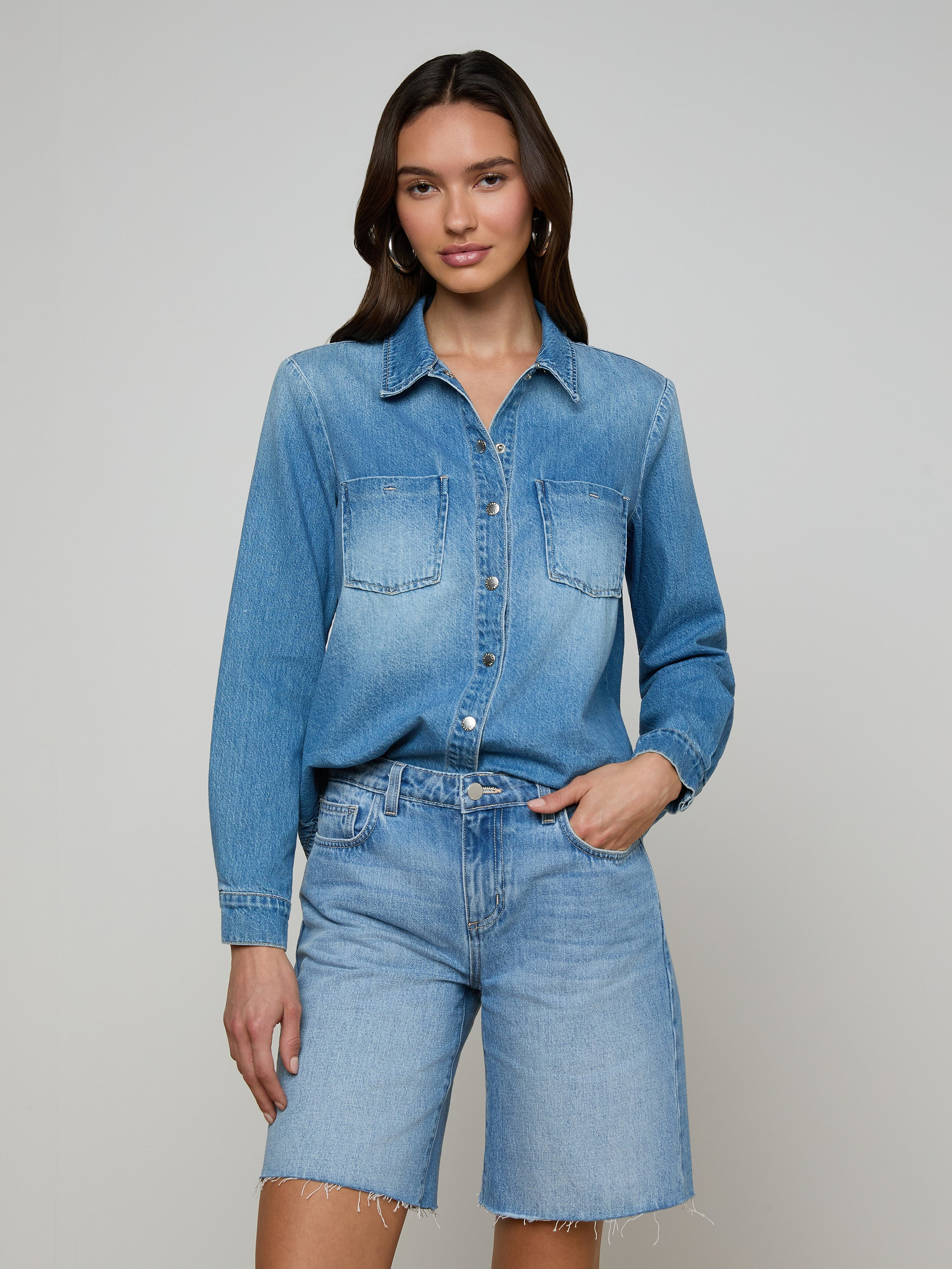 Wearing the Isla Shirt in a lightweight denim fabric and denim shorts, a woman stands against a plain light background with one hand in her pocket and a relaxed expression.