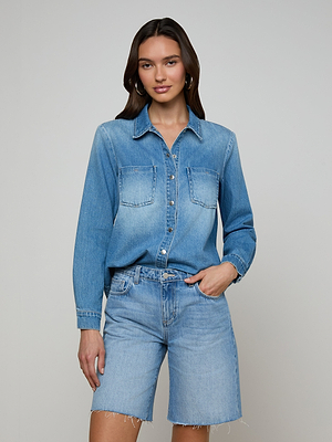 A woman with long dark hair and hoop earrings stands against a plain background wearing the Isla Shirt—an all-cotton, long-sleeve denim top with front pockets—and matching aged indigo knee-length shorts, one hand in her pocket.