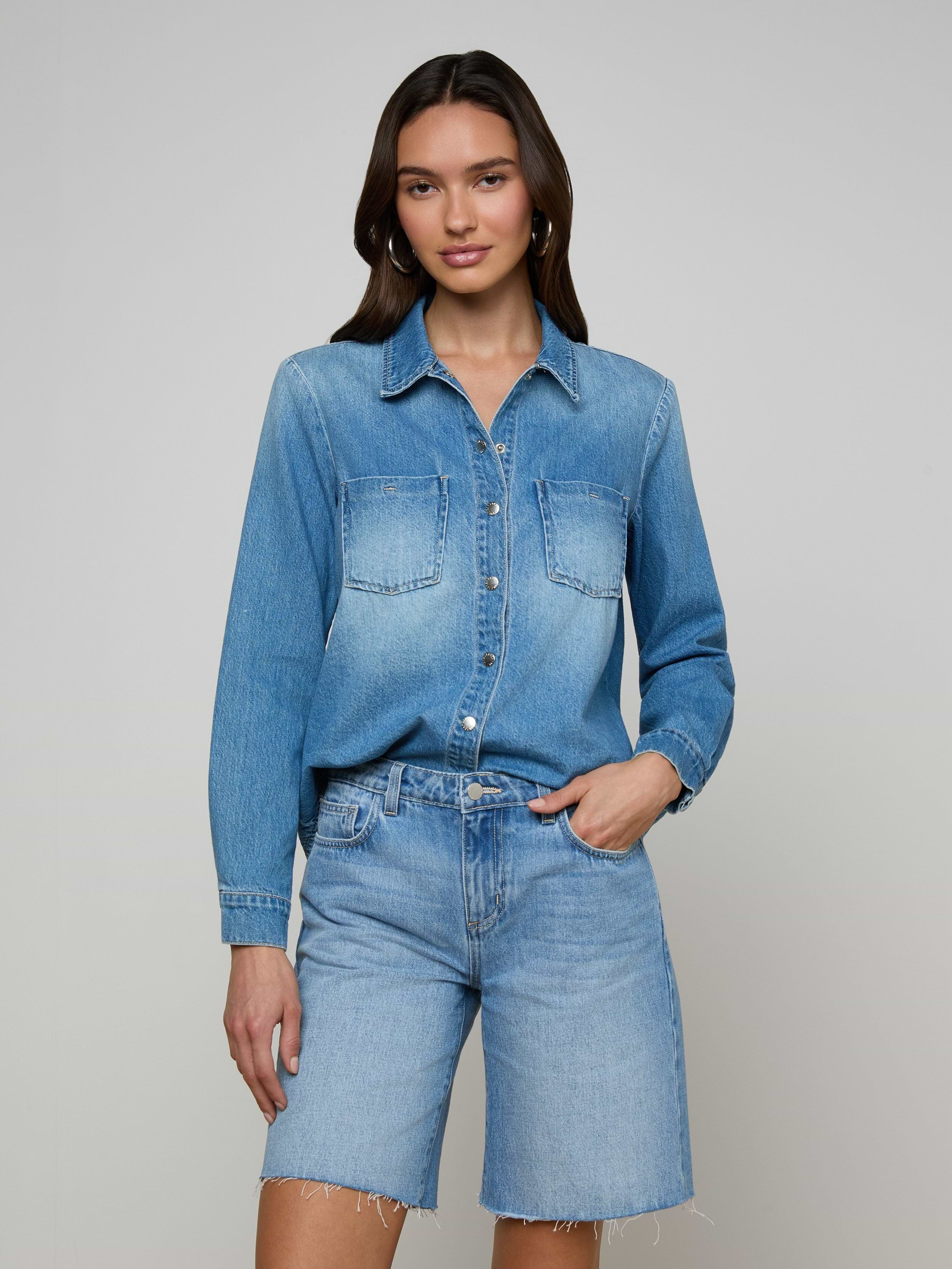 Wearing the Isla Shirt in a lightweight denim fabric and denim shorts, a woman stands against a plain light background with one hand in her pocket and a relaxed expression.