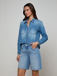 An individual stands against a plain, light background with one hand in her pocket, wearing the Isla Shirt—a lightweight denim shirt in an aged indigo wash—from L’AGENCE paired with matching knee-length denim shorts.