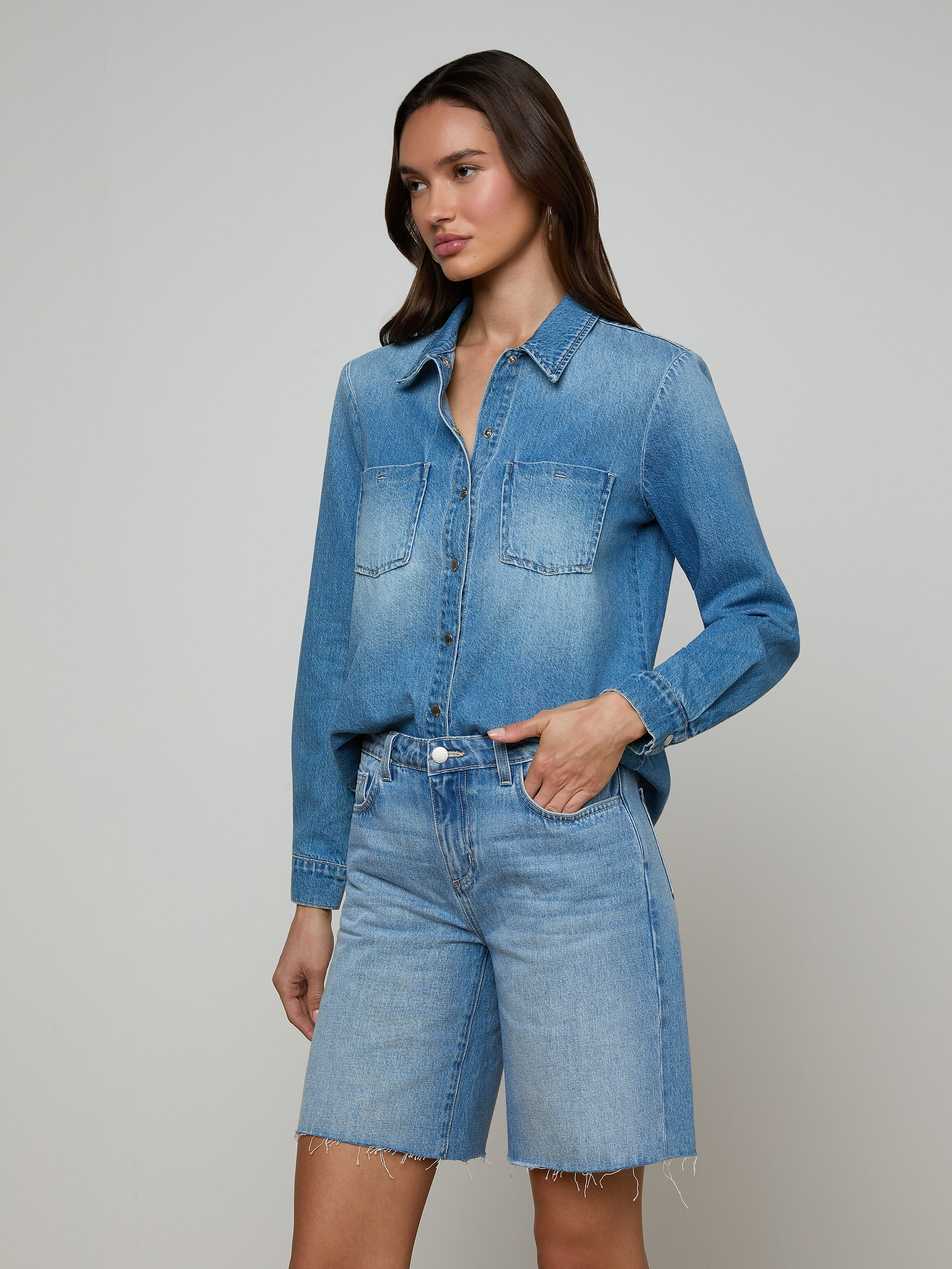 An individual stands against a plain, light background with one hand in her pocket, wearing the Isla Shirt—a lightweight denim shirt in an aged indigo wash—from L’AGENCE paired with matching knee-length denim shorts.