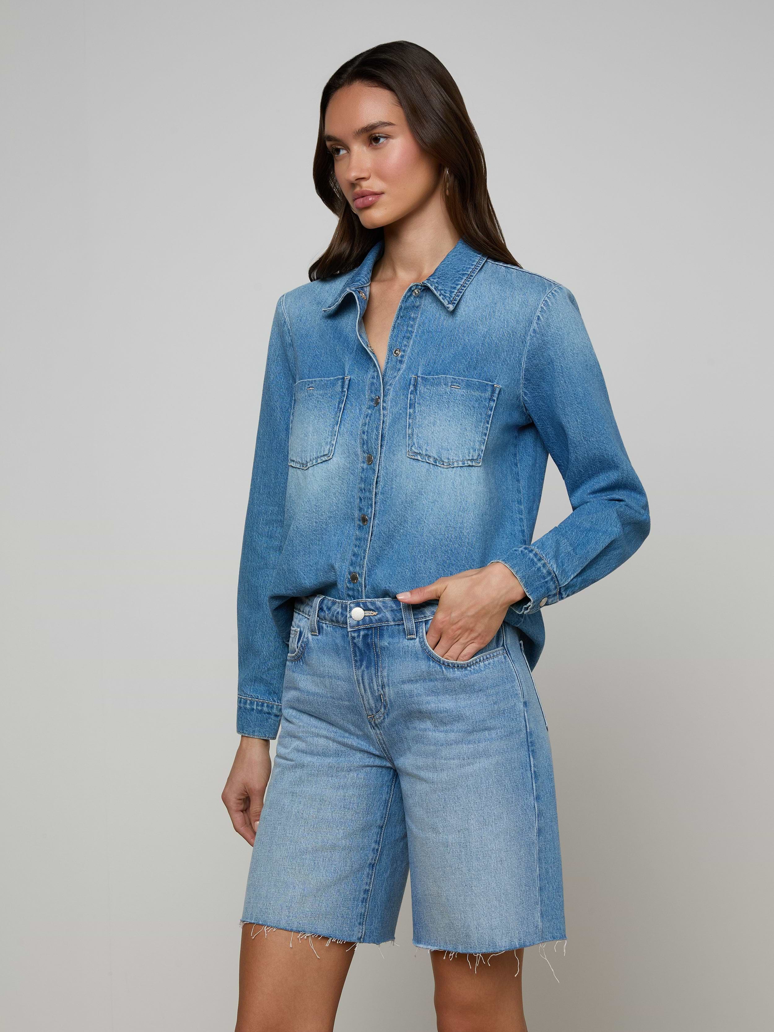An individual stands against a plain, light background with one hand in her pocket, wearing the Isla Shirt—a lightweight denim shirt in an aged indigo wash—from L’AGENCE paired with matching knee-length denim shorts.