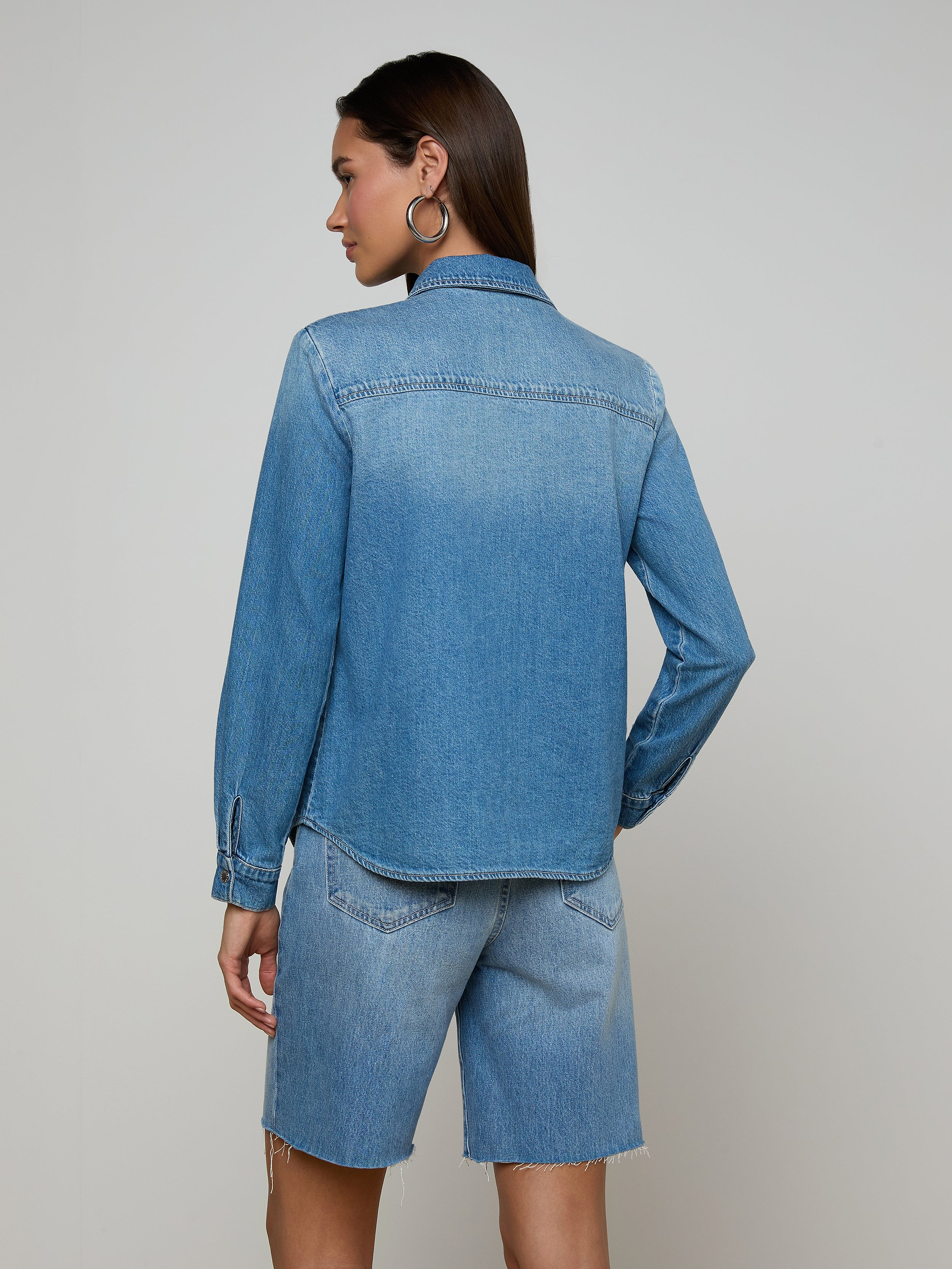 A woman stands with her back to the camera, wearing the lightweight Isla Shirt and matching denim shorts in an aged indigo wash, set against a plain off-white background.