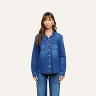 A woman with long brown hair stands facing forward, wearing the Isla Shirt—a featherweight denim shirt with front pockets—and matching blue jeans, against a plain light background.