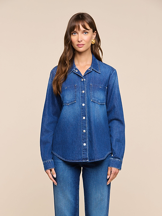 A woman stands facing forward wearing the Isla Shirt, a lightweight denim top in a medium-dark wash, paired with matching blue jeans against a plain beige background.