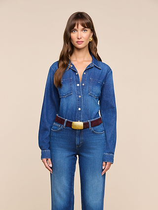 A woman with long brown hair wears the Isla Shirt, a lightweight denim top, paired with medium-dark wash jeans as she stands against a plain beige background, looking directly at the camera with a neutral expression.