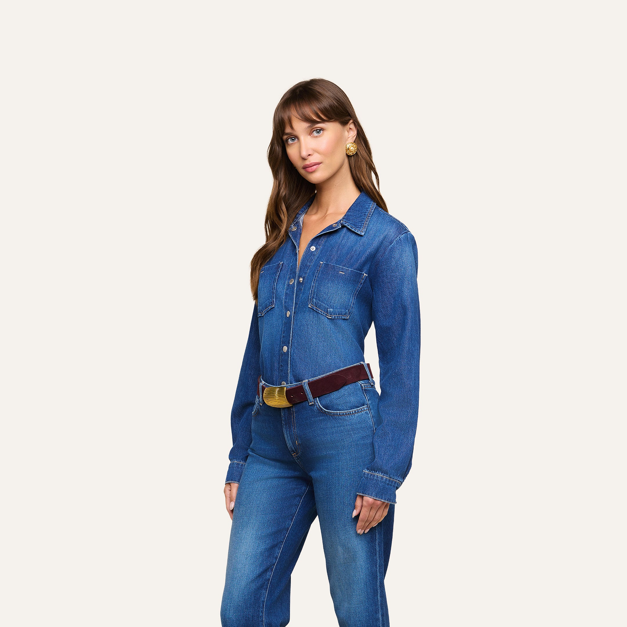 A woman wearing the Isla Shirt tucked into high-waisted jeans with a brown belt stands against a plain white background. She has long brown hair and is looking at the camera.