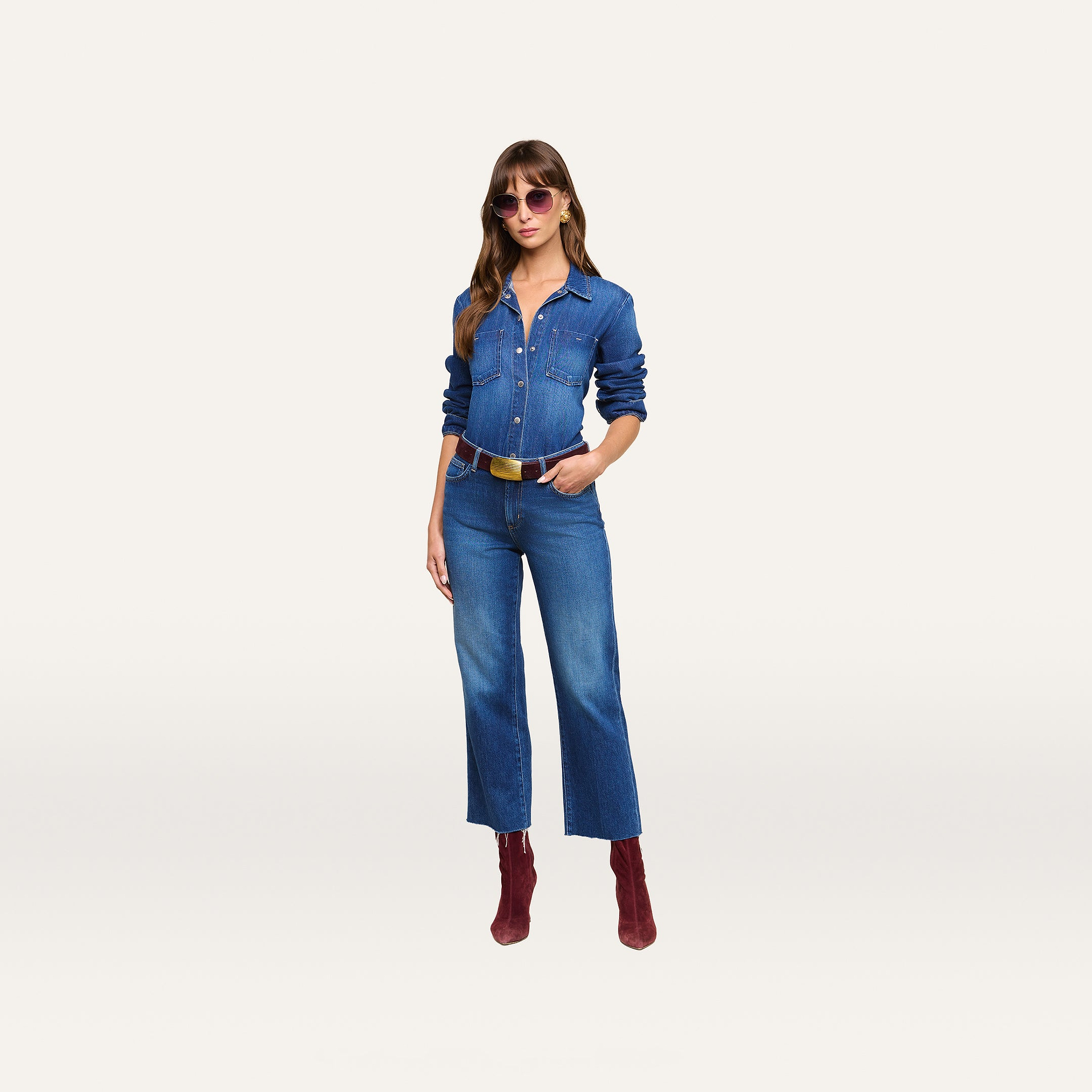 Woman in sunglasses wearing the Isla Shirt styled as a featherweight denim jumpsuit, paired with red ankle boots and a belt, standing with one hand in her pocket against a plain white background.