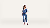 A woman wearing sunglasses, a blue denim shirt, blue denim jeans, a brown belt, and red boots stands against a plain white background.