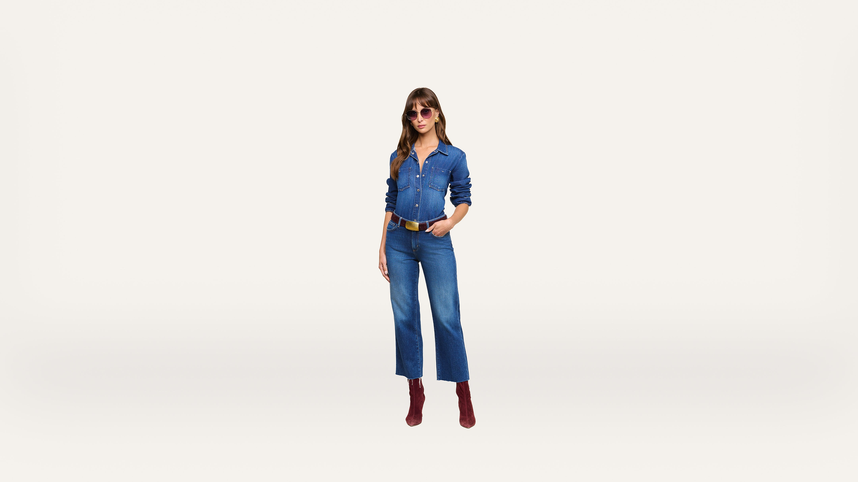A woman wearing sunglasses, a blue denim shirt, blue denim jeans, a brown belt, and red boots stands against a plain white background.