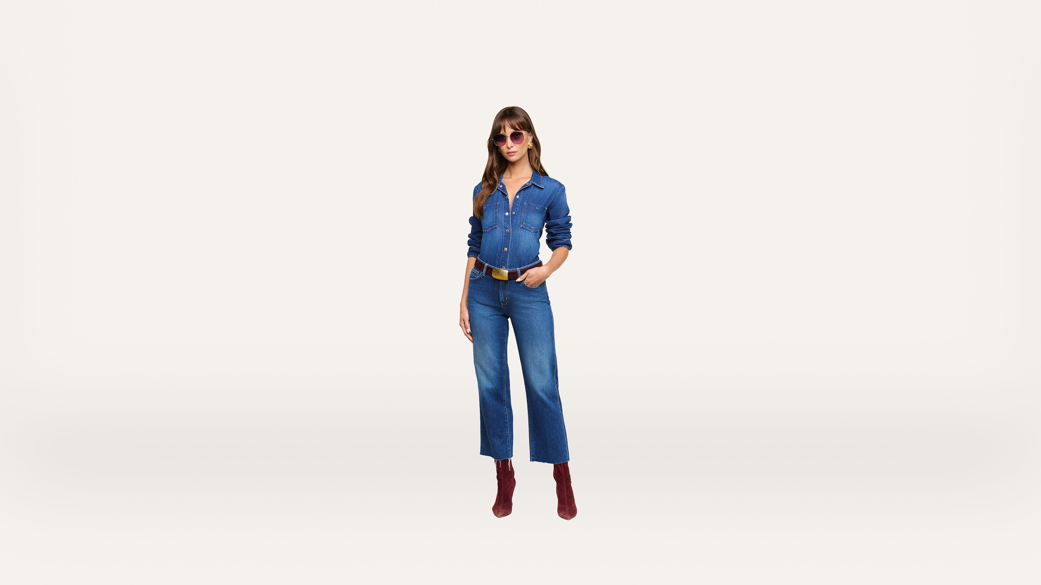 A woman wearing sunglasses, a blue denim shirt, blue denim jeans, a brown belt, and red boots stands against a plain white background.