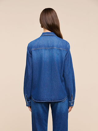 A woman with long brown hair stands facing away, wearing the Isla Shirt in featherweight denim and matching medium-dark wash denim pants against a plain beige background.