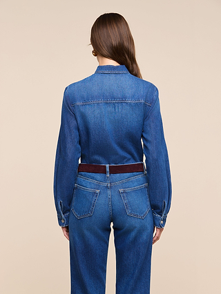 A woman with long brown hair stands facing away, wearing the Isla Shirt—a featherweight denim shirt—paired with medium-dark wash jeans and a dark belt, against a plain beige background.