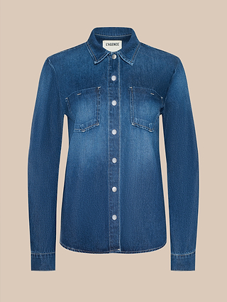 The Isla Shirt is a lightweight denim button-up in a medium-dark blue wash, featuring long sleeves, front chest pockets, and white buttons, shown against a plain beige background.