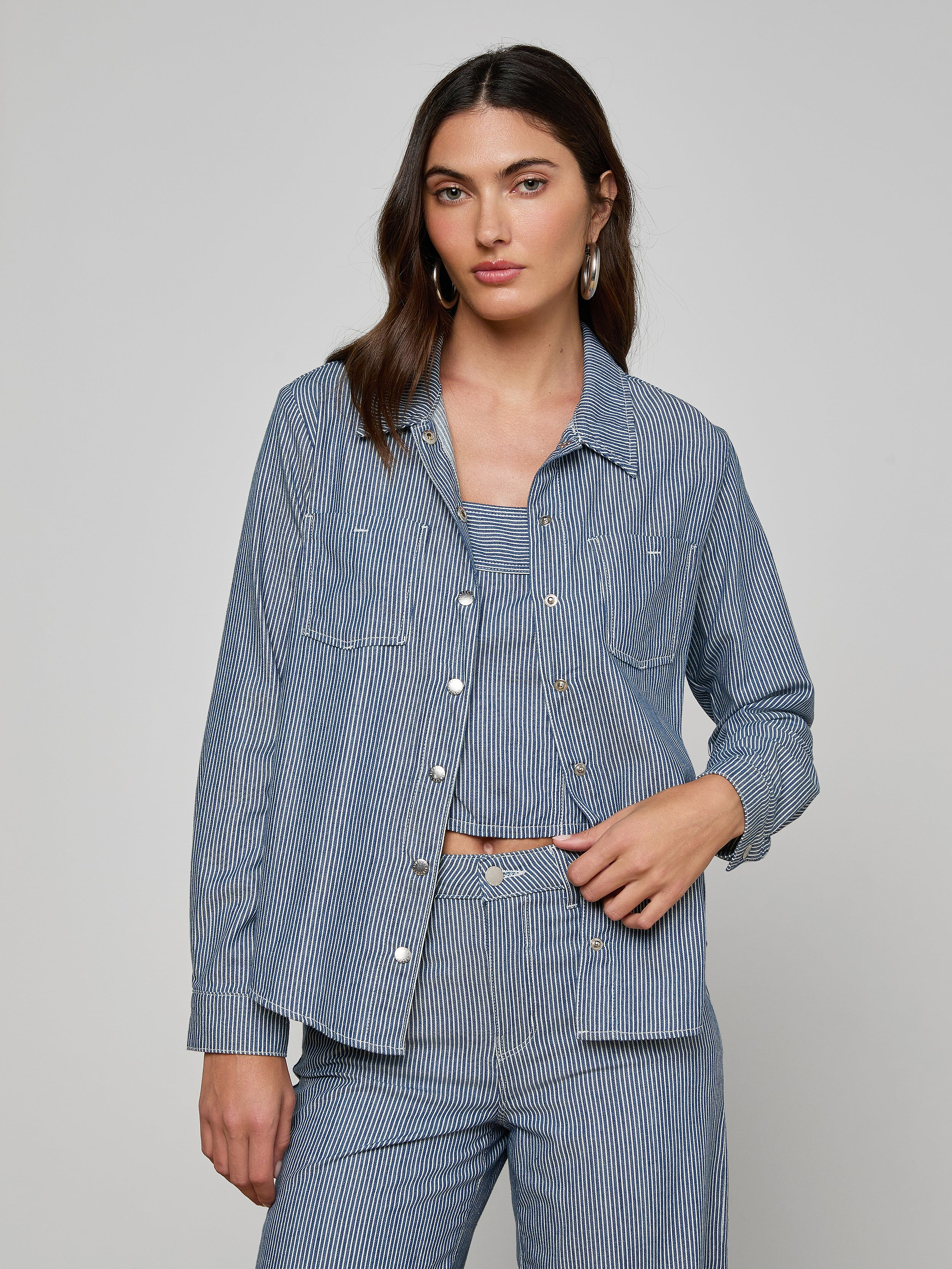 A woman wearing the Isla Shirt by L’AGENCE in featherweight indigo pinstripe denim over a coordinating top and pants stands against a plain light background, looking at the camera with a relaxed expression.