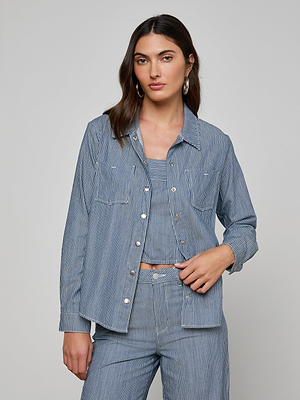 A woman with long brown hair wears the Isla Shirt by LAGENCE, an indigo pinstripe featherweight denim top, over matching striped pieces. She stands against a plain light gray background with a neutral expression and one hand on her hip.
