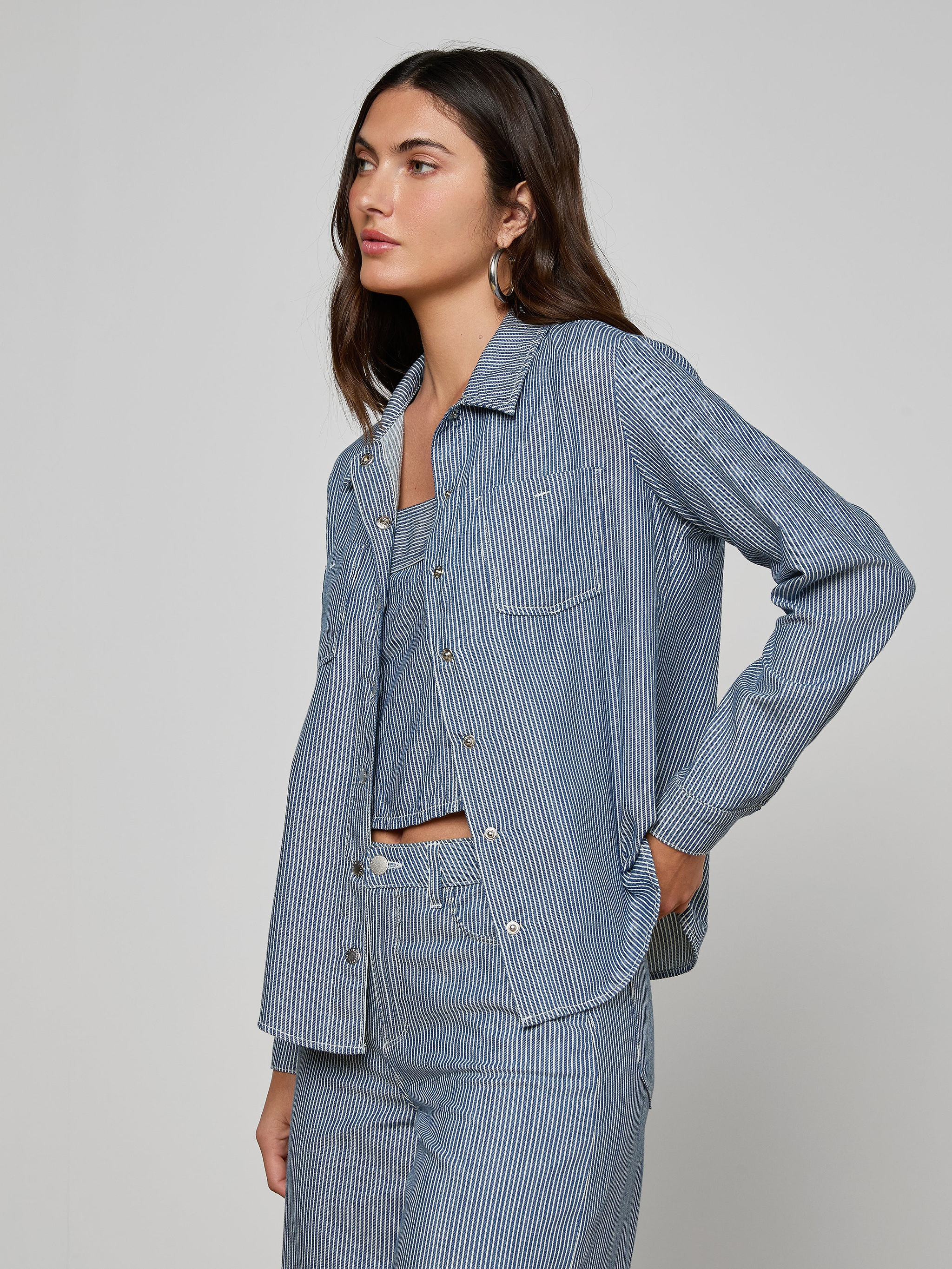 A woman stands against a plain background wearing the Isla Shirt in indigo pinstripe with matching pants, her left hand in her pocket and a neutral expression.