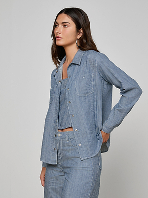 Woman with long brown hair wears the Isla Shirt in indigo pinstripe featherweight denim from LAGENCE, paired with matching pants, standing with one hand in her pocket and looking to the side against a plain background.