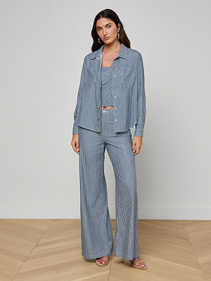 A woman stands indoors on a wooden floor, wearing the light blue Isla Shirt from L’AGENCE as part of an indigo pinstripe set with a crop top and wide-leg pants. She has long dark hair, a neutral expression, and is wearing sandals.