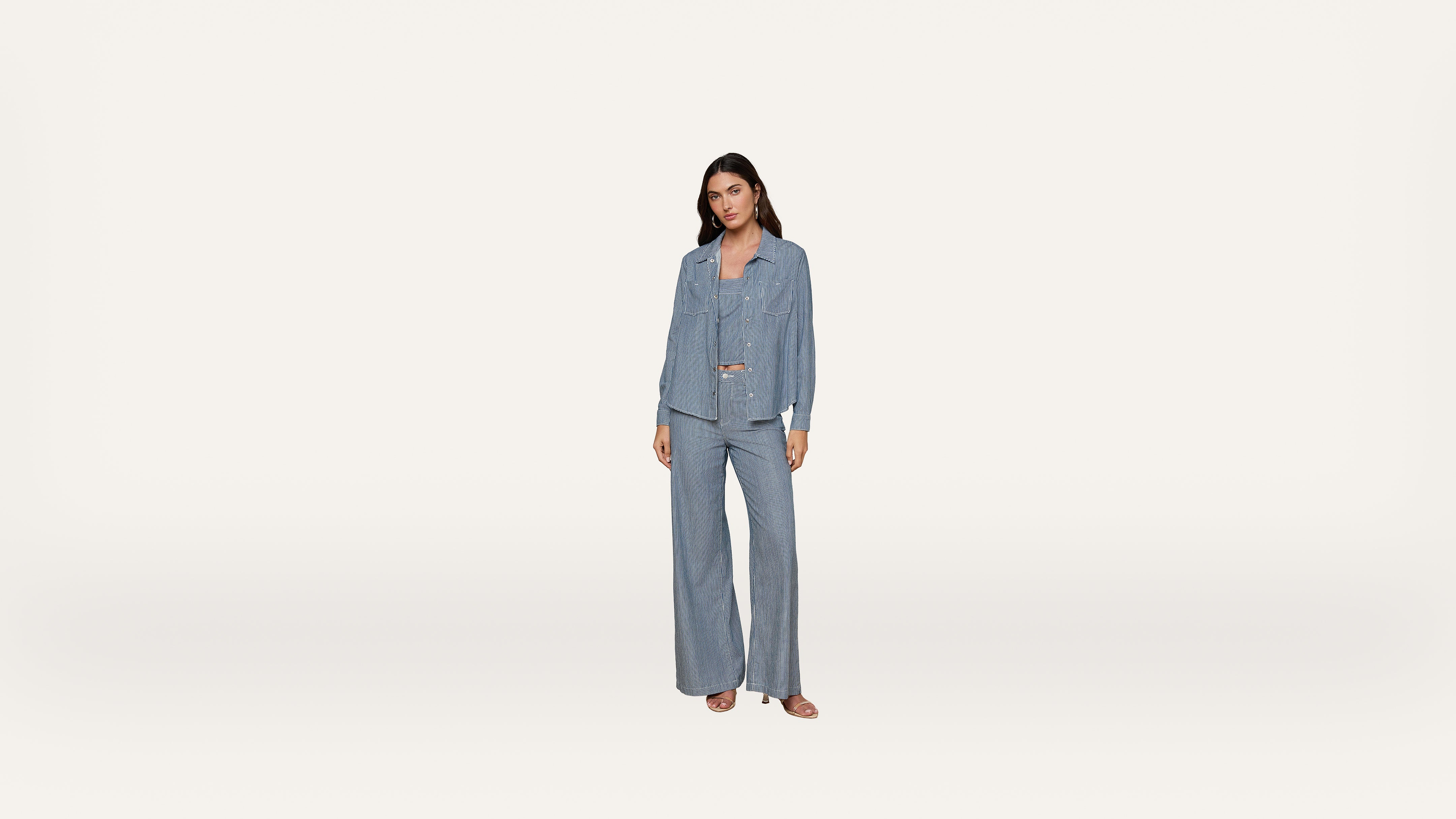 A woman stands wearing a matching light blue denim shirt and wide-leg jeans, paired with sandals, against a plain white background.