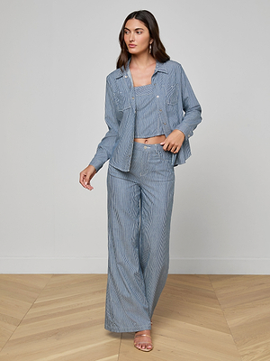 A woman stands on a wooden floor in an LAGENCE indigo pinstripe outfit: the Isla Shirt, a matching crop top, and wide-leg pants. She looks to the side, holding her shirt open with one hand.