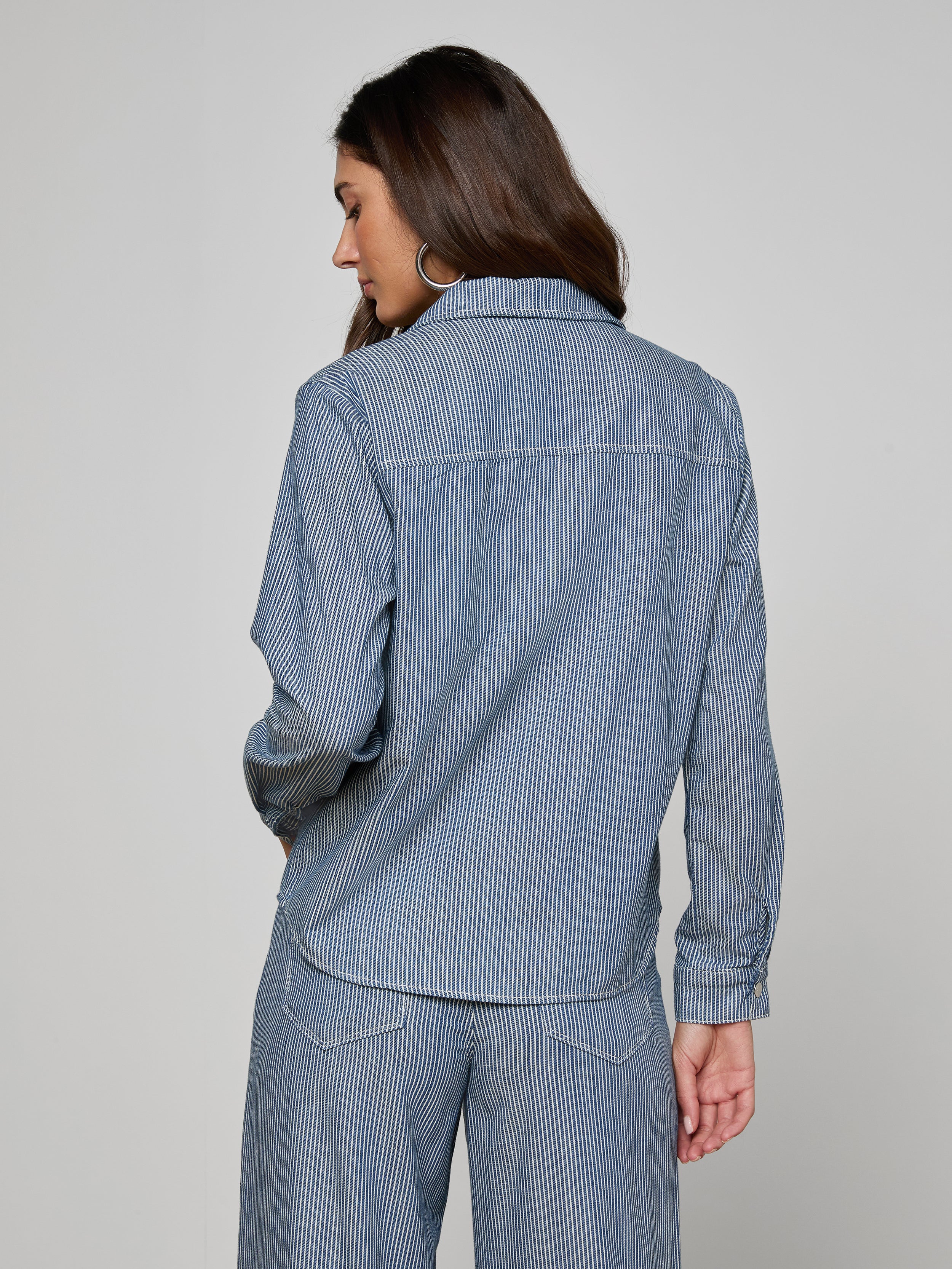 Woman standing with her back to the camera, wearing the Isla Shirt in indigo pinstripe featherweight denim from L’AGENCE, paired with matching pants, against a plain light background.