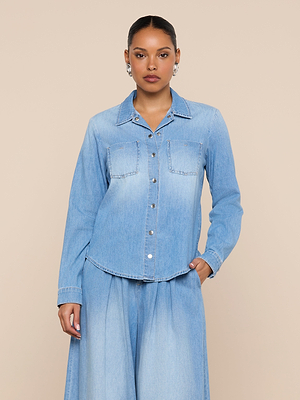 A person wearing the Isla Shirt, a lightweight vintage wash denim top with chest pockets, and matching wide-leg denim pants stands against a plain beige background.