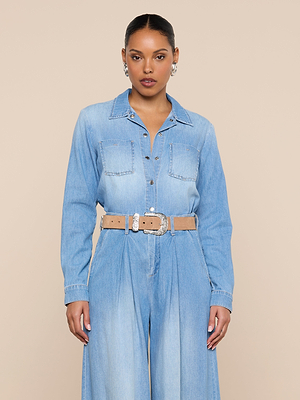 A woman stands facing forward in the Isla Shirt, styled with a beige belt and silver earrings, against a plain beige background.